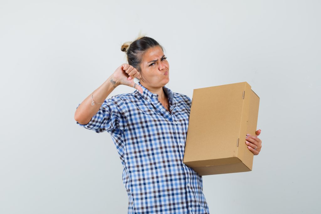 Young woman in checkered shirt holding box while showing thumb down and looking displeased , front view.